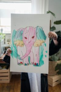 A child proudly displays a colorful elephant drawing indoors, showcasing creativity and art.